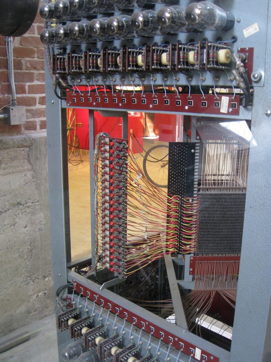 Photo of a tall gray rack from MIT's Whirlwind computer, about 1951, with vacuum tubes along the top and bundles of red and yellow wiring running to a core memory section.
